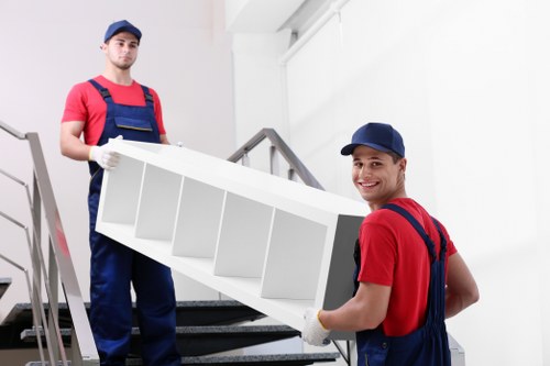 Two professional removal workers wearing red shirts, dark blue overalls, and matching blue caps are depicted in an indoor residential setting, likely a hallway or staircase landing in an Elephant and Castle property. The worker in the foreground is smiling and holding a large, white, wooden shelving unit with multiple compartments, positioned to be carried down a staircase. The shelving unit appears to have a smooth, painted finish. The second worker stands behind him on the staircase, holding the other side of the shelving unit, with a serious expression. The background features a white wall and a metallic handrail, indicating the interior of a typical London terraced house or flat. The environment is well-lit with natural or artificial light, emphasizing the professional nature of the removals service provided by Elephant and Castle Man with Van, focused on careful handling of household furniture during the moving process within a residential area in south London postcode.