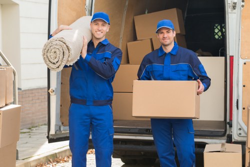 Two professional removalists dressed in blue uniforms and matching caps stand in front of an open removals van on a residential driveway in London, with a house visible in the background. One removalist on the left is smiling and holding a rolled-up rug with a woven texture, while the other on the right is also smiling and carrying a medium-sized cardboard box. The van's interior is filled with various packed cardboard boxes, some sealed and others open, as well as large pieces of furniture partially visible inside. The scene depicts a house move in progress, with the removalists ready to transport household items such as furniture, boxes, and personal belongings, demonstrating careful handling and organization typical of local house removals in the Elephant and Castle area. The lighting is natural, suggesting daytime, and the setting emphasizes professional and efficient removal services aligned with the company's offerings in central London postcodes.