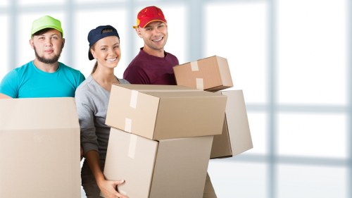 The image depicts three professional movers standing indoors in front of a bright, airy residential setting with large windows that let in natural light. The movers are dressed in casual work attire, with two wearing baseball caps, one red and one navy, and the other wearing a light green cap. They are smiling and appear to be in good spirits as they hold several cardboard boxes of varying sizes, which are sealed with packing tape and have a typical brown finish. The boxes are made of sturdy cardboard with some showing packing tape reinforcement along edges, indicating they are prepared for transportation. The interior environment suggests a home, with a smooth, light-colored wall visible behind them. The scene emphasizes careful handling and organization typical of house removals, supported subtly by the use of boxes and the professional demeanor of the movers, which aligns with the relocation services offered by Elephant and Castle Man with Van in the London area, possibly the South London postcode that includes Elephant and Castle.