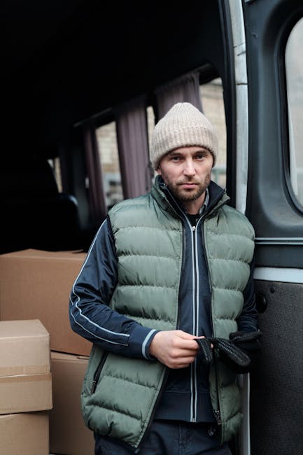 A man standing beside a moving van during a home relocation, dressed in a light grey knitted beanie, a green padded vest over a dark blue jacket with white piping along the sleeves, and black gloves.