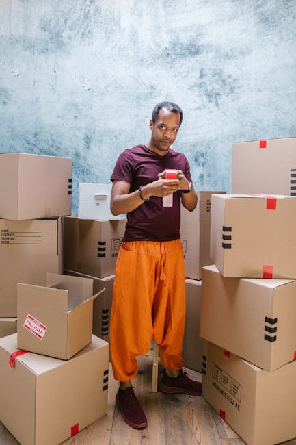 A man inside a room, surrounded by multiple cardboard moving boxes of various sizes, some sealed with red tape and others open revealing packing materials. The boxes are stacked on the floor and on a