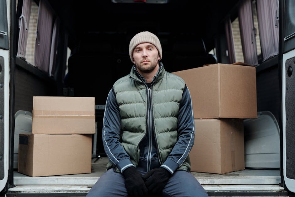 A man wearing a beige knit beanie, black gloves, a blue and green puffer jacket, and dark pants is seated on the pavement in front of a partially loaded van. The interior of the van contains several c