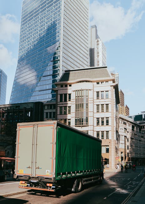 A man wearing a beige knit beanie, black gloves, a blue and green puffer jacket, and dark pants is seated on the pavement in front of a partially loaded van. The interior of the van contains several c